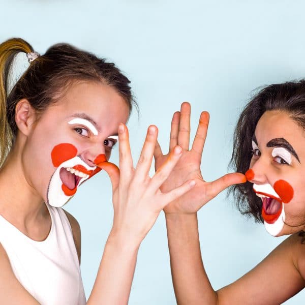 Two teenage brunettes in t-shirts and with painted faces jokingly show their long noses with their fingers palm. Isolated on gray. Family celebrates April Fool’s Day.